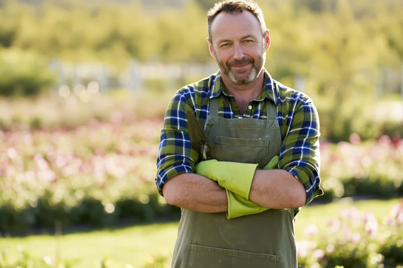 Smiling man in apron and gloves standing in garden with arms crossed